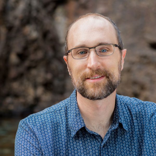A picture of Ben Adams in front of a rock wall that is blurred behind him in the background. He is a white bearded man with brown short hair wearing glasses and smiling. He is in a short-sleeved button up blue shit with white swirls and yellow dots.